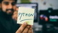 A man of color sitting next to a few computers holding a post it that says "Python" in the foreground. 